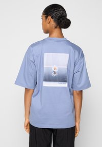 Jasnoniebieski, oversize bawełniany t-shirt z prostokątnym kwiatowym motywem na plecach, z napisem "Calvin Klein" na dole.