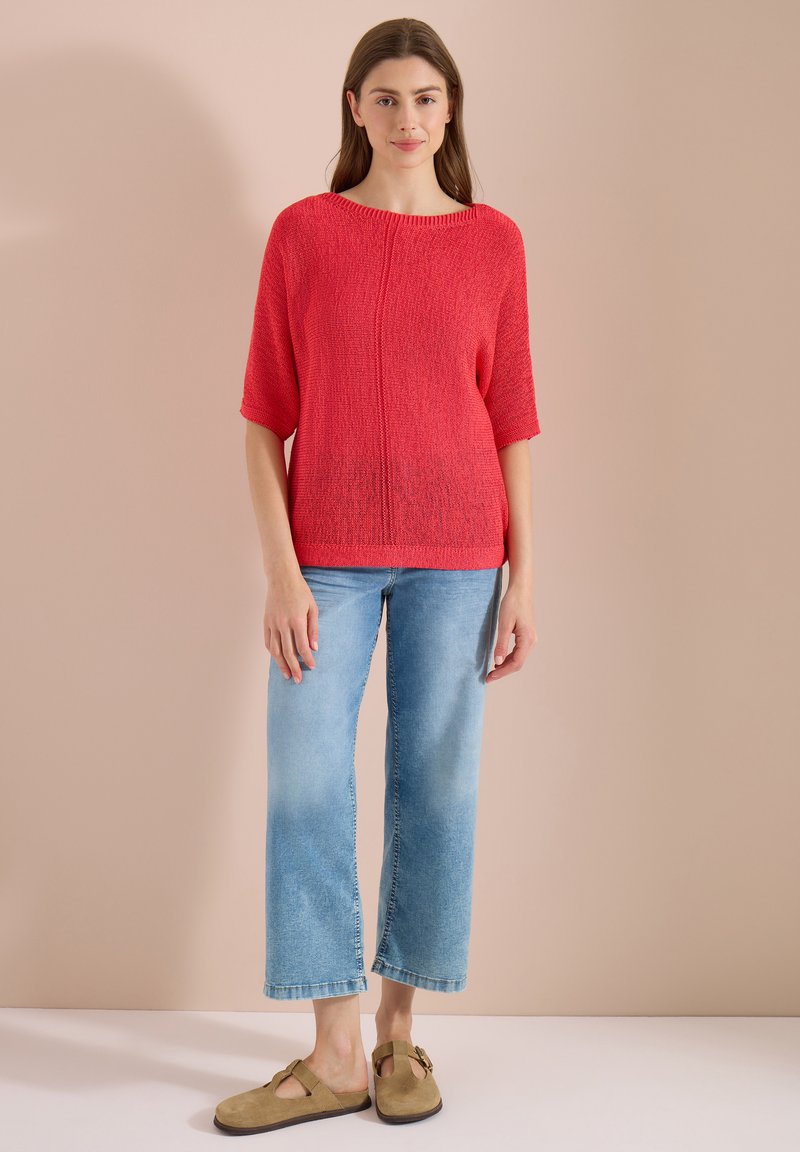Femme debout portant un pull rouge à manches courtes, un jean court bleu clair et des chaussures marron sans lacets sur un fond beige.