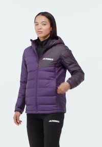 Kobieta w fioletowo-ciemnoszarej kurtce puchowej Terrex z kapturem, zapinana na zamek, w połączeniu z czarnymi spodniami Terrex na białym tle.