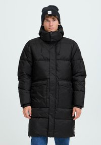 Melna puffa jaka ar augstu apkakli, pogām un sānu kabatām. Izgatavota no kviltes materiāla. Valkāta virs zilām džinsām. Melna beanie cepure.