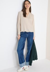 Haut beige à manches longues avec un col en V, associé à un jean denim bleu clair. Le jean présente une coupe décontractée et des ourlets effilochés. Baskets blanches avec des accents rouges.