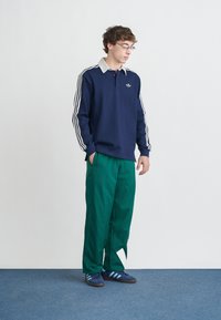 Tengerészkék hosszú ujjú póló, fehér gallérral és három fehér csíkkal az ujjakon, zöld nadrággal és csíkos sötétkék tornacipővel párosítva.