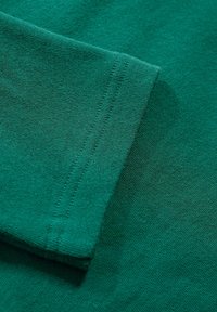 Groene katoenstof met een gladde textuur; close-up van een mouw die stikdetails en een omgeslagen rand laat zien.