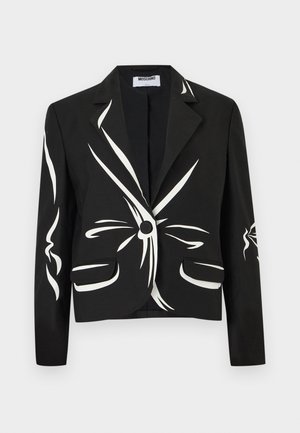 Femeie purtând un sacou negru cu linii albe abstracte, pantaloni negri cu crăpături largi și încălțăminte neagră cu degetele descoperite, stând pe un fundal alb simplu.