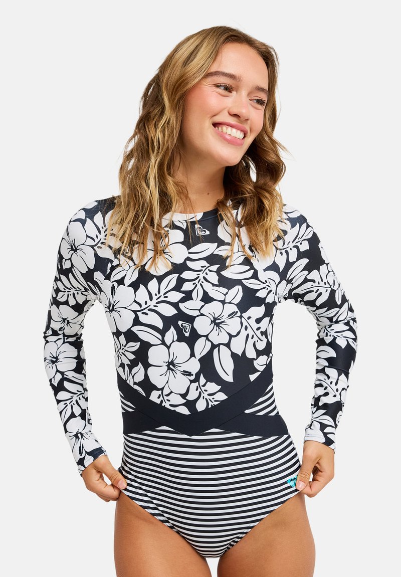 Νεαρή γυναίκα χαμογελά, φορώντας μαγιό με μακριά μανίκια, μαύρο-λευκό floral τοπ και ριγωτό κάτω μέρος πάνω σε απλό φόντο.