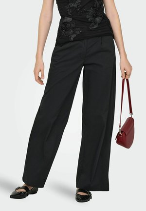 Donna con pantaloni neri a gamba larga e top nero a fiori, che tiene una piccola borsa rossa, in piedi con i piedi divaricati e scarpe nere piatte.