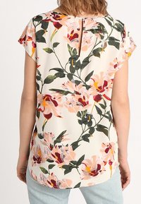 Donna che indossa una blusa a maniche corte con una grande stampa floreale in rosa e bordeaux, con chiusura a bottoni a chiave sul retro, abbinata a jeans azzurri.