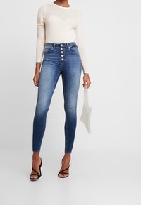 Jeans de mezclilla azul de cintura alta con una textura suave, que cuentan con cinco botones y un desvanecimiento degradado. Combinados con una blusa de manga larga de canalé color crema y un bolso transparente.