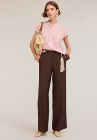 Blusa rosa chiaro a maniche corte con bottoni, abbinata a pantaloni larghi marrone scuro con tasche laterali e una superficie texturizzata. Scarpe e borsa beige.