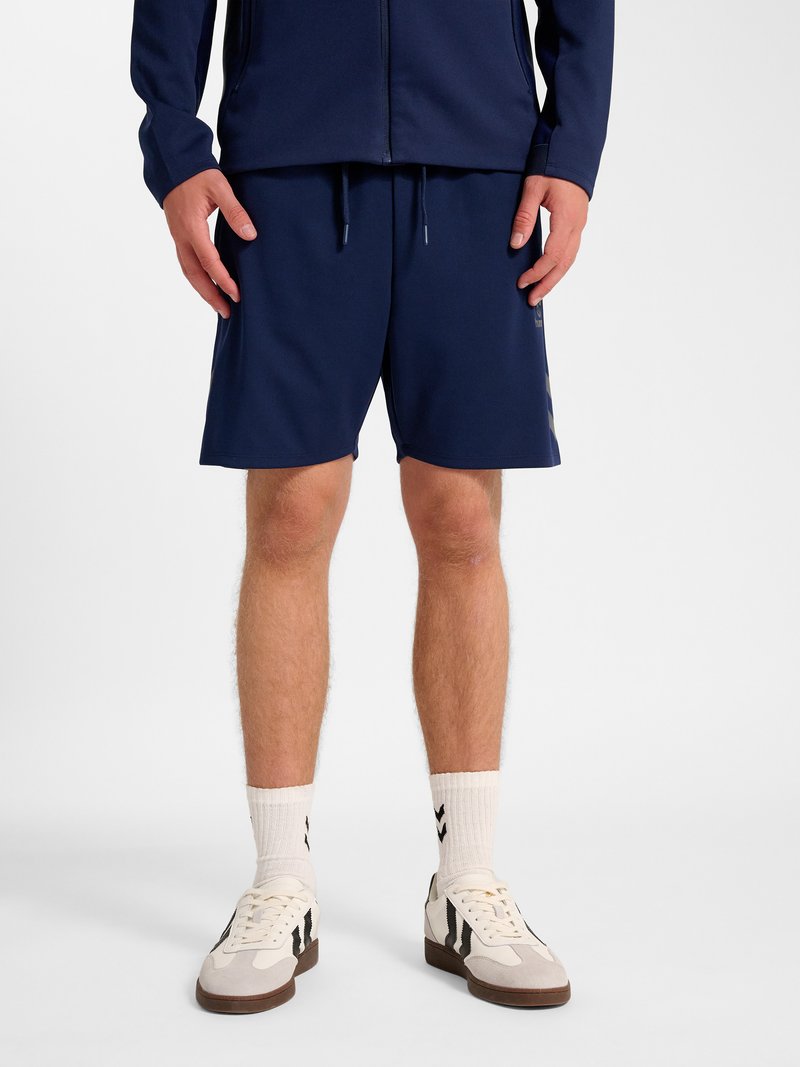 Man draagt marineblauwe korte broek, marineblauw jasje, witte sokken en witte sneakers met zwarte strepen, staand tegen een effen achtergrond.