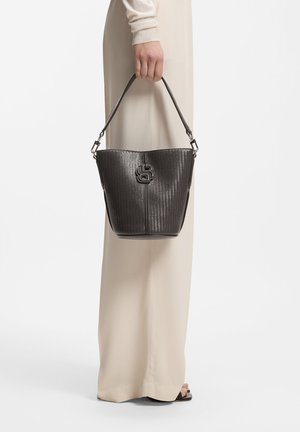 Person som har på seg beige bukser og sandaler, som holder en svart teksturert bøtteveske med kort håndtak og metallogo-detalj.