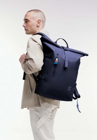 Zaino navy a chiusura arrotolata con spallacci regolabili, realizzato in materiale resistente. Presenta una tasca frontale con zip e tirazip colorati.