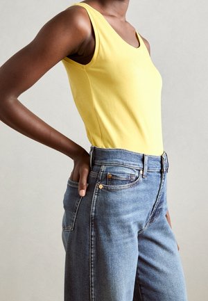 Person trägt ein ärmelloses gelbes Oberteil, das in eine hoch taillierte blaue Jeans gesteckt ist, und hat eine Hand auf der Hüfte.