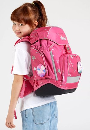 Junges Mädchen mit roten Haaren und Pferdeschwanz, trägt ein weißes Hemd und blaue Jeans, hält einen großen rosa Rucksack mit Sternenmuster und Einhorn-Ansteckern.