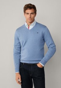 Maglione blu chiaro a V realizzato in materiale morbido, con un logo discreto sul petto e polsini a coste, abbinato a jeans scuri.