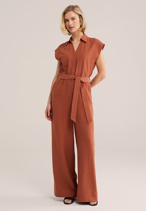 Nainen seisoo osittain käsin selän takana, yllään ruosteenvärinen, ohuilla raidoilla kuvioitu, hihaton, leveälahkeinen haalari jossa vyö solmittuna, mustat sandaalit.