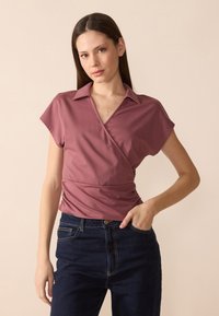 Wrap-style topp i støvet rosa med kragesnitt, korte ermer og rynkedetaljer i midjen, paret med mørk denim jeans.
