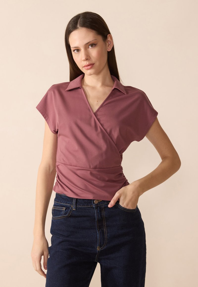 Top stile wrap in rosa polveroso con colletto, maniche corte e dettagli arricciati in vita, abbinato a jeans in denim scuro.