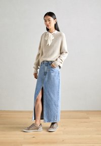 Pull en maille beige à col cravaté, associé à une jupe maxi en denim bleu clair avec une fente sur le côté. Des baskets beiges complètent le look.