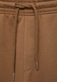 Pantaloni de trening maro cu închidere prin șnur, talie ribbată și textură netedă cu vârfuri de cordon argintii. Buzunar lateral vizibil.
