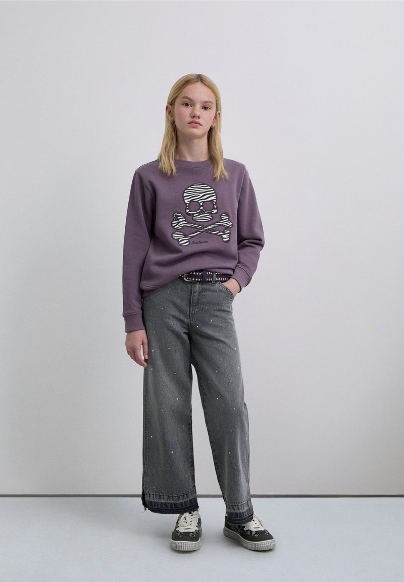 Sweatshirt violet avec un motif tête de mort et os croisés, associé à un jean gris délavé à jambes larges et déchiré avec des embellissements en perles et des baskets noires.
