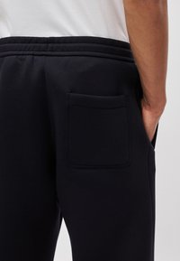 Schwarze Jogginghosen mit flachem Bund und einer einzelnen Tasche auf der Rückseite, aus weichem Stoff mit glatter Textur.