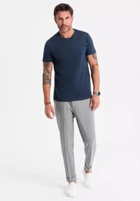 T-shirt blu navy a maniche corte abbinato a pantaloni grigi chiari plissettati e scarpe slip-on bianche. Presenta tatuaggi sul braccio sinistro.