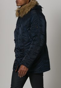 Giacca puffer blu navy con cappuccio bordato di pelliccia, chiusure a zip e bottoni. Presenta una tasca laterale con zip e superficie in tessuto testurizzato.