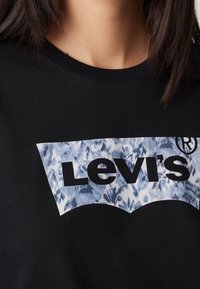 Svart bomullströja med en stor Levi's-logotyp i fet svart text över en blå och vit blommig grafik på bröstet.