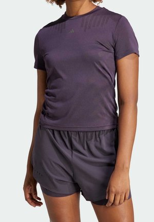 Personne portant un t-shirt de sport moulant violet foncé et un short assorti, debout sur un fond clair uni.