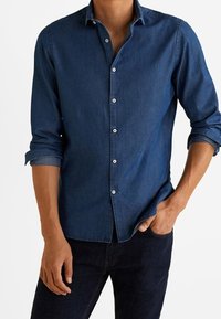 Uomo che indossa una camicia di jeans scura a maniche lunghe con le maniche arrotolate e pantaloni scuri, con una mano in tasca, in piedi contro uno sfondo bianco.