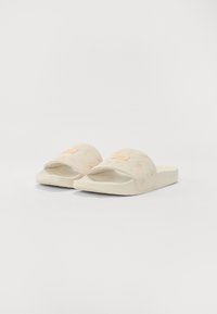Des sandales beige clair avec un motif floral texturé sur la bride, présentant un accent logo orange à l'avant et une semelle plate épaisse.