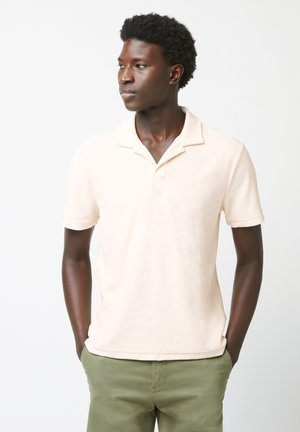 Jeune homme aux cheveux courts et bouclés portant un polo à manches courtes beige clair et un pantalon vert, les mains dans les poches, regardant vers la gauche.