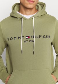 Felpa con cappuccio verde oliva realizzata in materiale morbido, con tasca frontale, coulisse bianche e logo blu navy con accenti rossi e bianchi.