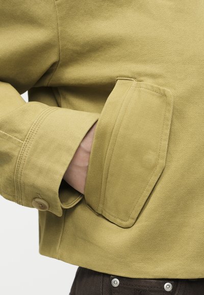 Χέρι μερικώς εισαχθέν στην πλαϊνή τσέπη ενός κίτρινο-πράσινου μπουφάν με μανσέτα με κουμπιά και ορατό πτερύγιο τσέπης.