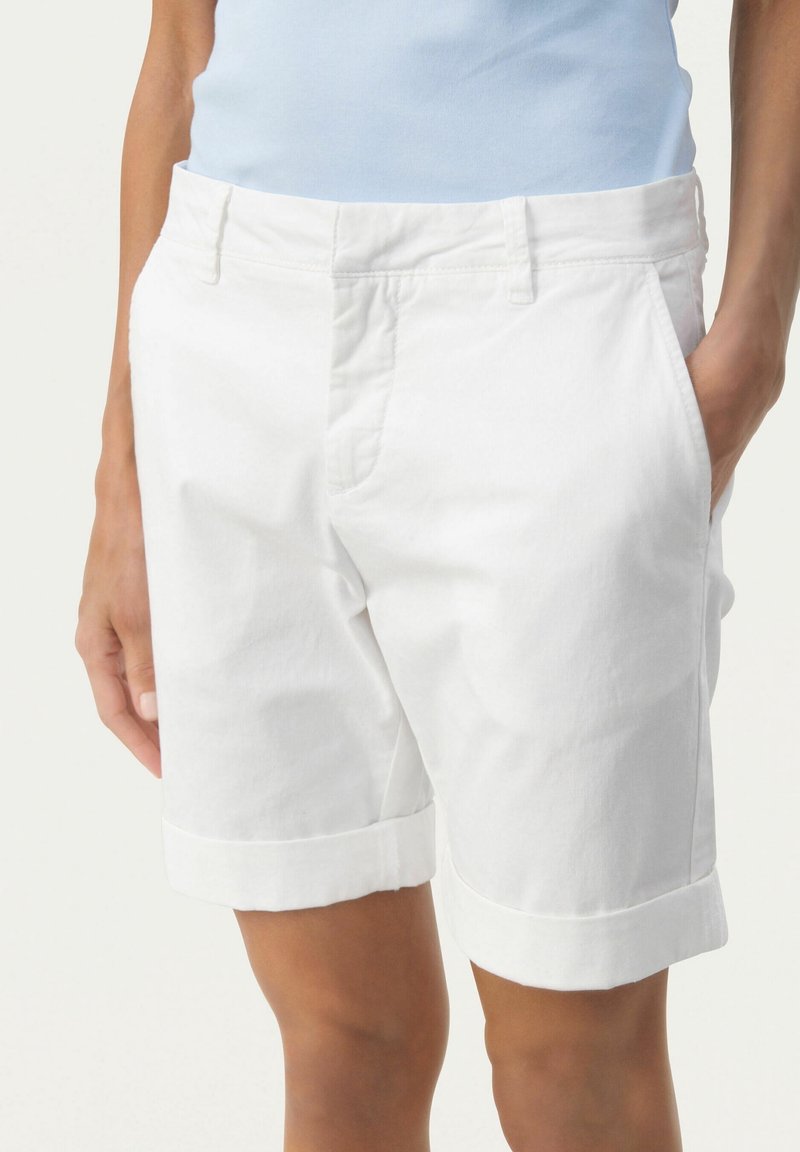 Personne portant un short blanc arrivant aux genoux avec des ourlets retournés et des poches latérales, associé à un haut sans manches bleu clair.