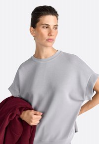 Helles graues T-Shirt mit kurzen Ärmeln in lässiger Passform, mit einem gerippten Halsausschnitt. Das Modell hält einen burgunderfarbenen Pullover, der über ihrem Arm hängt.