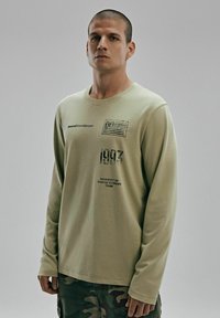 Jasnozielona koszula z długim rękawem z nadrukowanymi grafikami, w tym designem flagi, rokiem "1993" i tekstem, zestawiona z spodniami w kamuflażu.