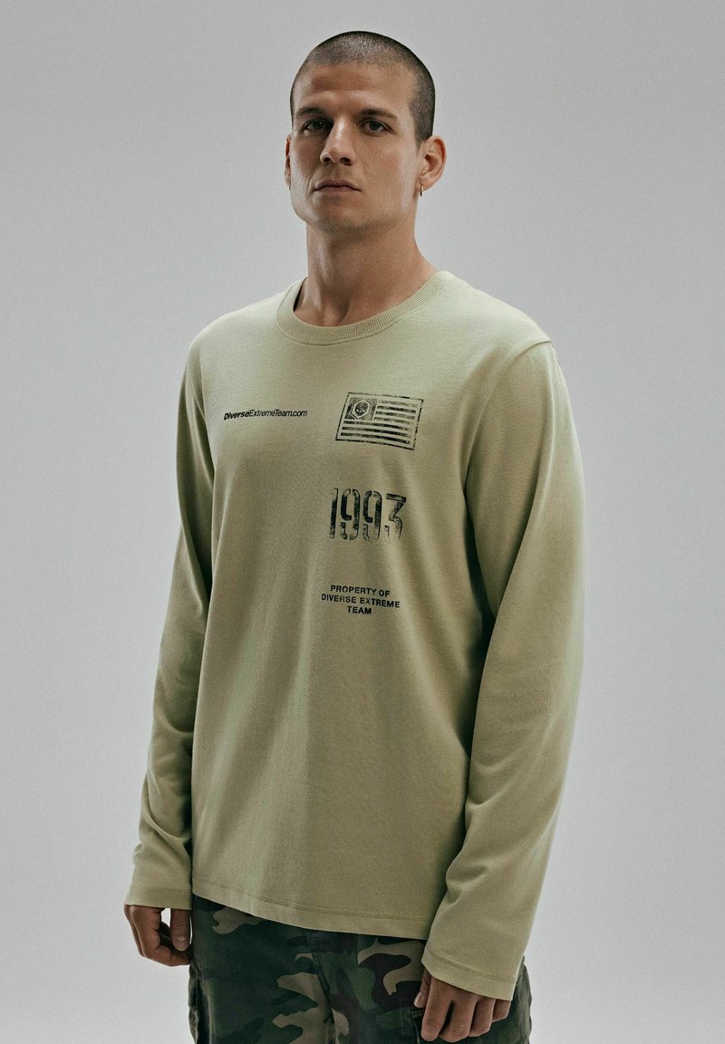 Jasnozielona koszula z długim rękawem z nadrukowanymi grafikami, w tym designem flagi, rokiem "1993" i tekstem, zestawiona z spodniami w kamuflażu.