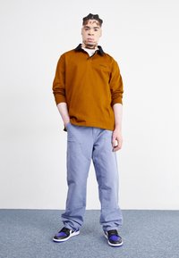 Hnědá polo košile s dlouhým rukávem a černým límečkem, spárovaná s lehkými modrými volnými kalhotami se zlatými doplňky a černofialovými teniskami.