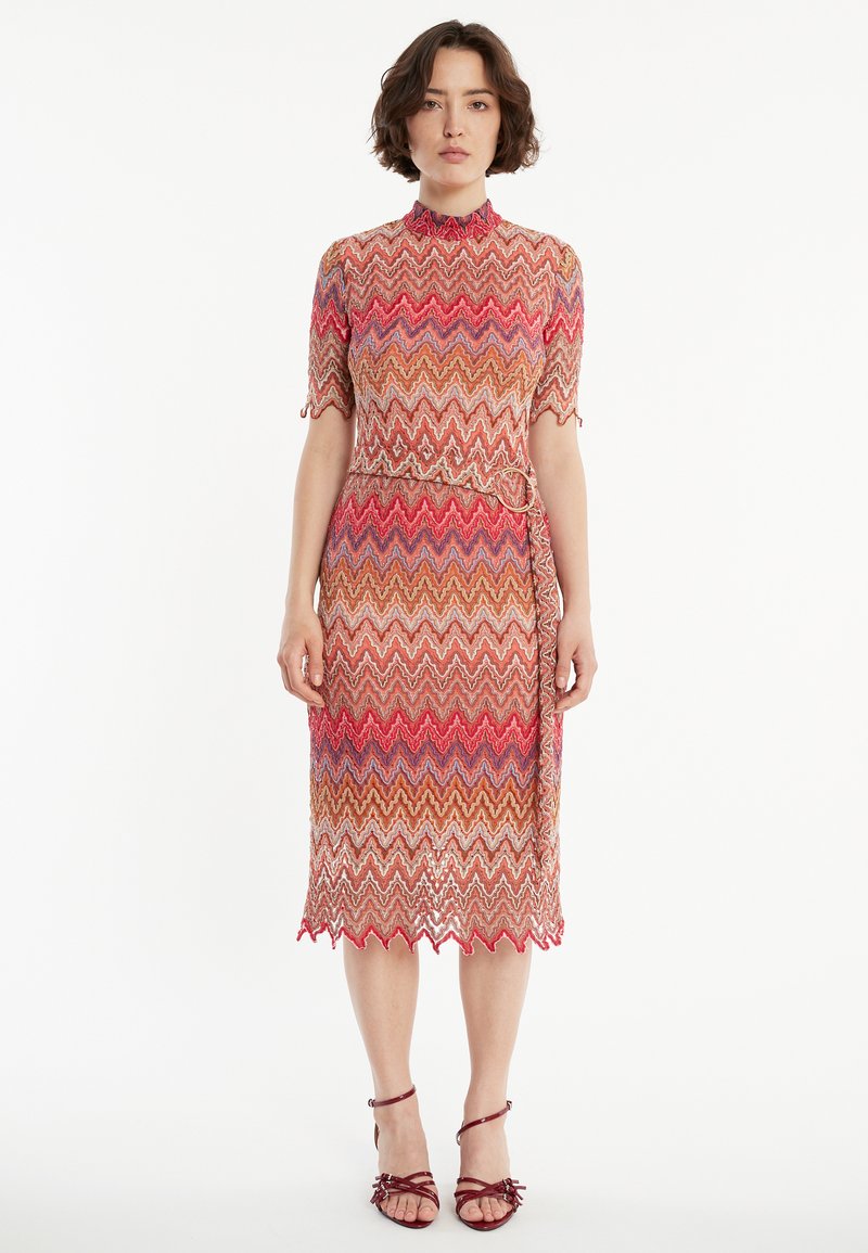 Knielanges, tailliertes Kleid mit einem Zickzackmuster in Rot-, Orange- und Beigetönen. Hat einen hohen Kragen und kurze Ärmel sowie einen Bindegürtel.