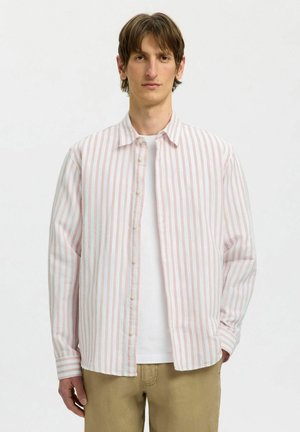 Młody mężczyzna ubrany w różowo-białą koszulę w pionowe paski na guziki, założoną na białą koszulkę, oraz beżowe spodnie, stoi na jednolitym tle.