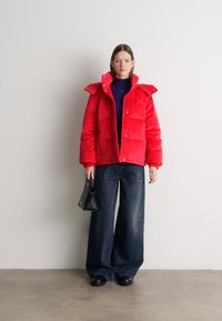 Sarkans pūkains mētelis ar redzamu quiltēto dizainu, augstu apkakli un spiedpogu aizdari, apvienots ar platām kājām tumšajām džinsa biksēm un mazu melnu rokassomu.