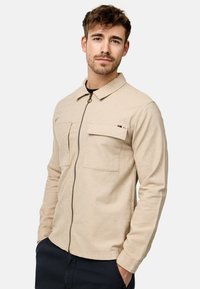 Veste beige à fermeture éclair à l'avant avec un col, deux poches poitrine et un tissu texturé. Présente un détail rayé subtil sur la poche.