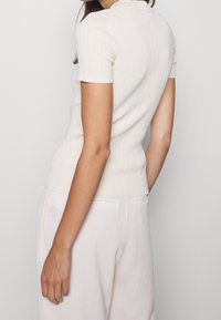 Top ajusté en tricot côtelé de couleur beige clair avec des manches courtes et un col polo. Texture lisse avec une coupe crop.