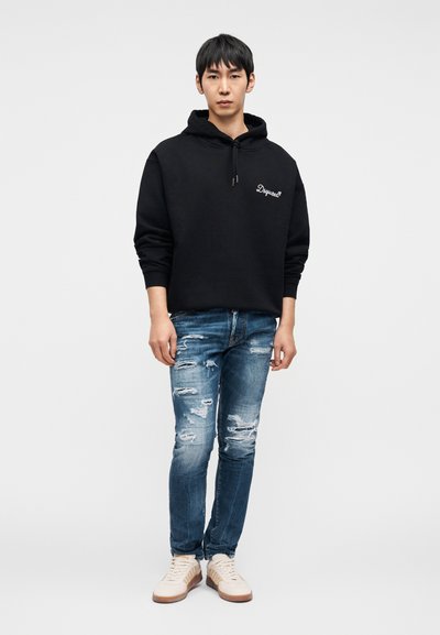 Μαύρο φούτερ με κουκούλα και λογότυπο μπροστά, συνδυασμένο με τζιν μπλε με φθορές και μπεζ αθλητικά παπούτσια με καφέ σόλες.