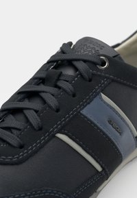 Sötétkék sneaker sima és struktúrált anyagok keverékével. Szürke részletekkel, textúrált cipőfűzőkkel és logóval a nyelvén.