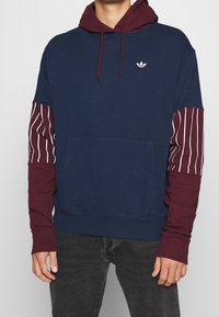 Huva sweatshirt i marinblått med vinröda ärmar som har vita vertikala ränder. Inkluderar en framficka och en liten logotyp på bröstet.