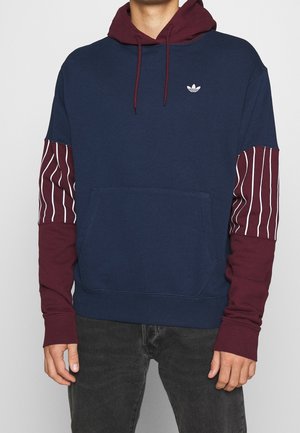 Mikina s kapucí v námořnické modři, s maroon rukávy a bílými vertikálními pruhy. Obsahuje přední kapsu a malé logo na hrudi.