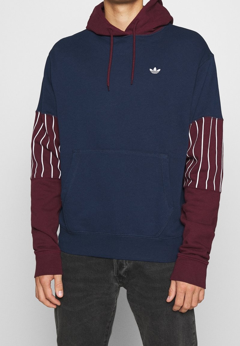 Huva sweatshirt i marinblått med vinröda ärmar som har vita vertikala ränder. Inkluderar en framficka och en liten logotyp på bröstet.